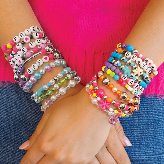 Two child's wrists wearing multiple colorful beaded bracelets, including letter beads spelling out words, pearl-like beads, metallic beads, and flower charms, set against a bright pink top and blue denim background.
