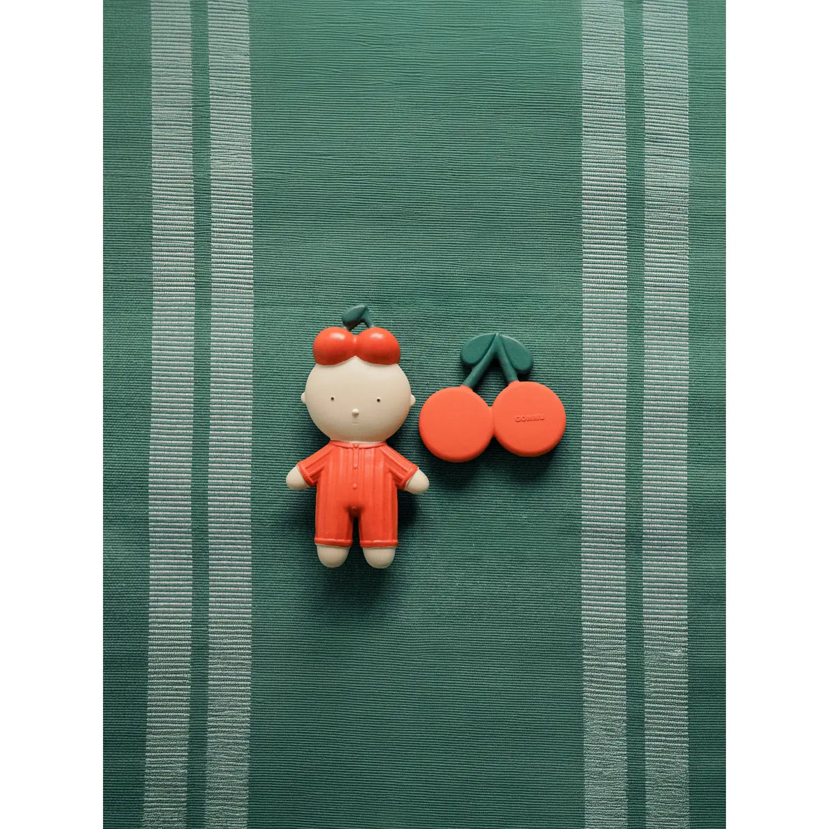 A small decorative figurine of a child in an orange outfit with red hair accessories, placed beside two orange cherries with green stems on a green striped fabric background.