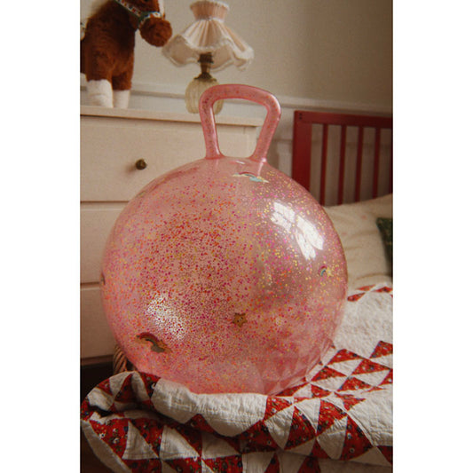 A translucent pink hopscotch ball with gold glitter speckles and a pink handle, resting on a red and white patterned quilt in a bedroom setting.