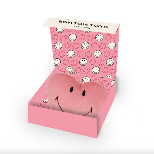 A pink plush heart-shaped toy with a smiling face, nestled inside an open pink gift box printed with smiley faces. The top flap of the box reads 'BON TON TOYS EST.