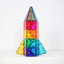 A colorful toy rocket built from transparent magnetic building tiles, standing upright on a white surface against a white paneled background.