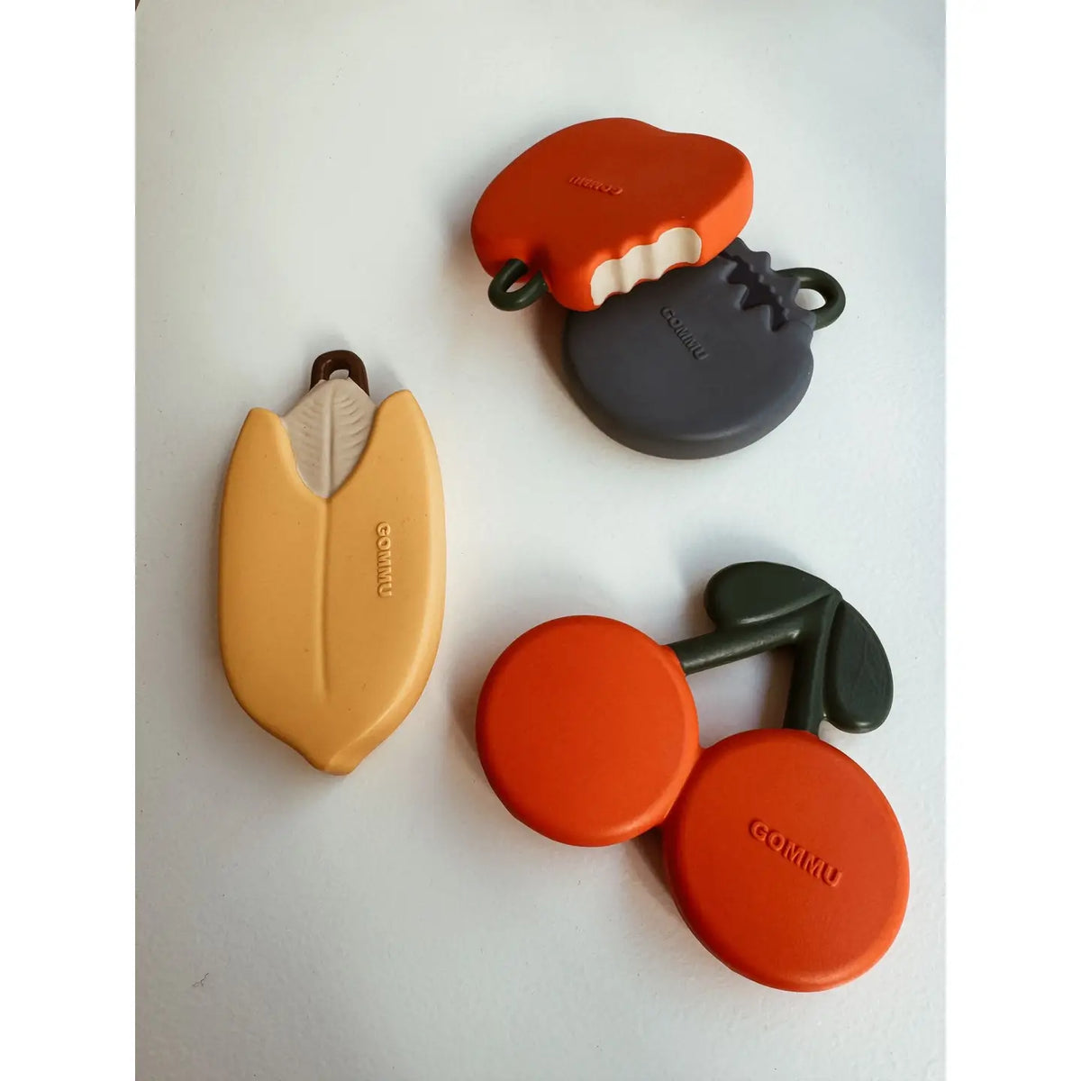 Four GOMMU silicone baby teething toys arranged on a light gray background: a yellow corn-shaped toy, an orange and dark gray tooth-shaped toy, and an orange and dark gray cherry-shaped toy.