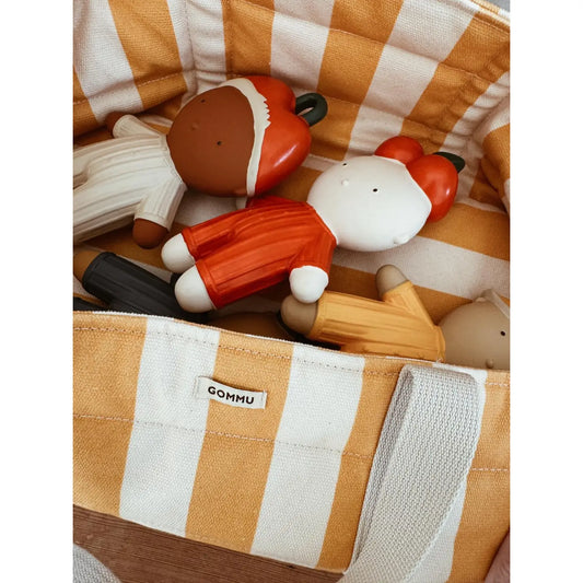 A close-up of a striped fabric storage basket or organizer containing wooden toy figures. The basket has alternating warm tan and off-white vertical stripes, with a small visible brand tag reading GOMMU.