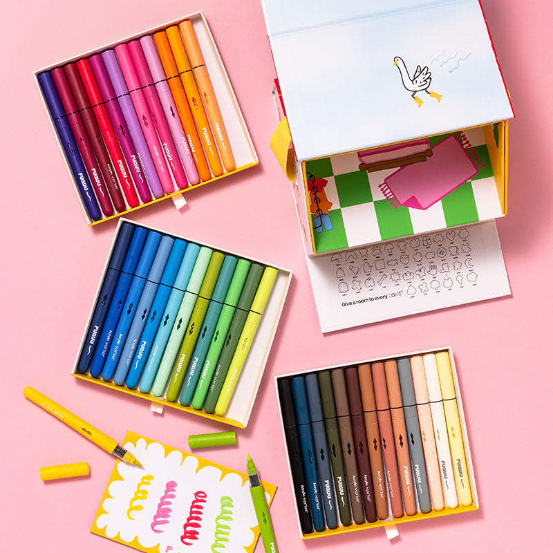 Four open boxes of acrylic pens arranged on a pink background, showing multiple color sets including reds, blues, greens, yellows, browns, and neutrals. One box is open to reveal a colouring book insert with a swan drawing, and crayons are scattered around the boxes.