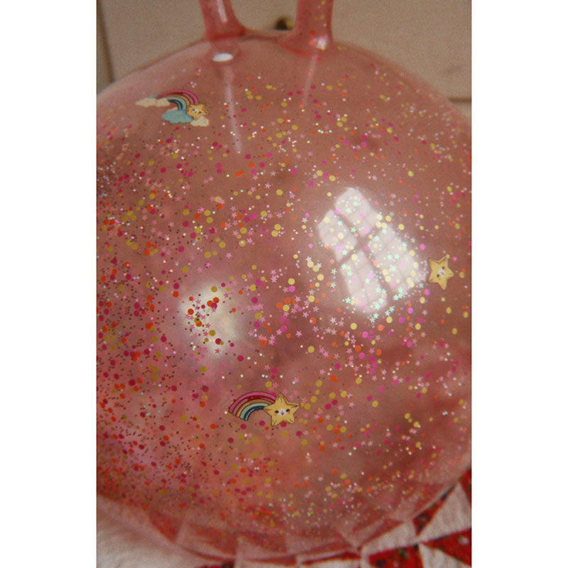 A translucent pink party balloon filled with multicolored glitter and decorated with small rainbow and star stickers, shown against a neutral indoor background.
