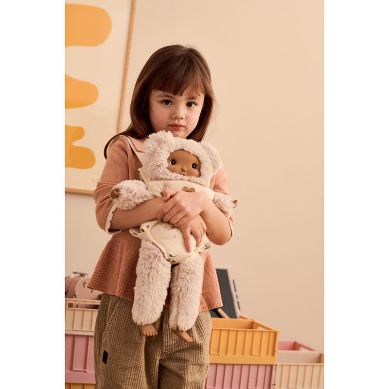 A young child wearing a dusty rose top and brown pants holds a plush doll dressed in a cream-colored sheep costume in a warm, softly lit room.