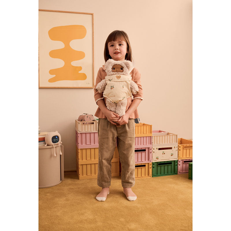 A young child stands in a softly styled nursery-like room holding a plush teddy bear dressed in a cream-colored bunny suit. The child wears a peach top and brown pants, with pastel storage crates and a framed abstract print in the background.