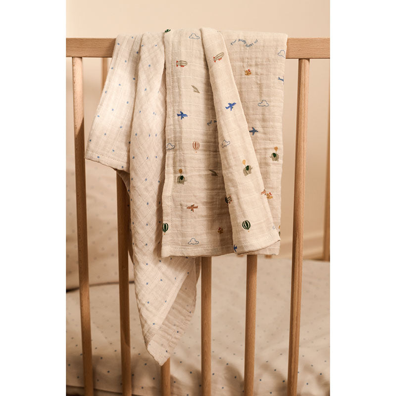A light beige muslin baby blanket with small embroidered motifs of airplanes, hot air balloons, and clouds, draped over the side of a wooden crib.