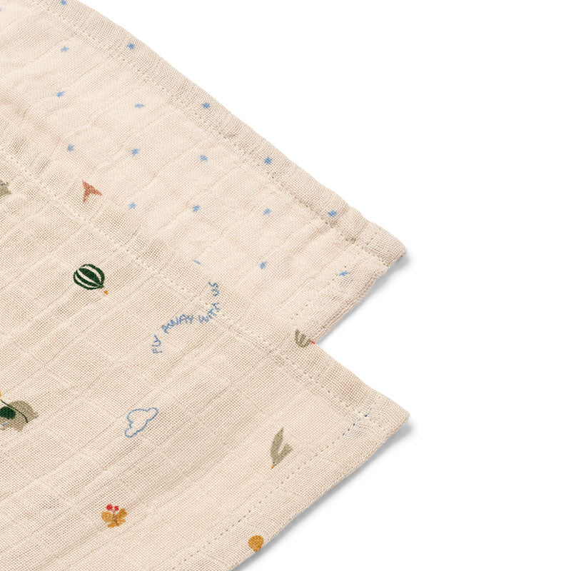 A close-up of two folded muslin baby cloths  in a warm cream colour, featuring a printed pattern of small blue stars, a green watermelon, a cloud, and tiny colorful motifs, with the words 'my happy baby' embroidered in blue.