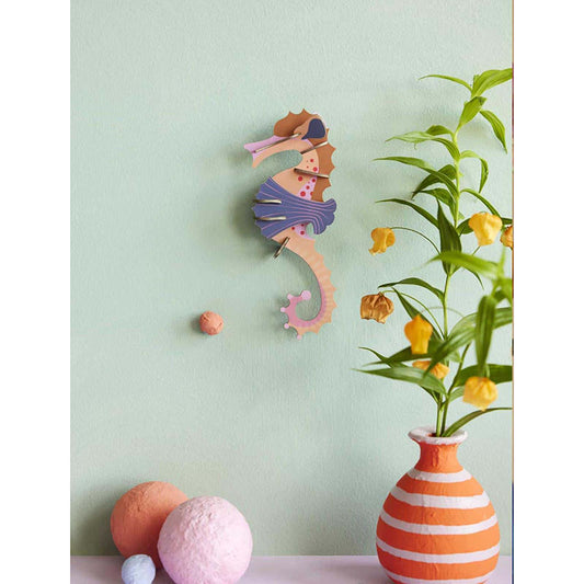 A decorative seahorse wall hanging in pastel pink, purple, and orange tones, displayed on a pale mint wall above a surface with a striped orange-and-white vase, yellow flowers, and soft pastel spheres.