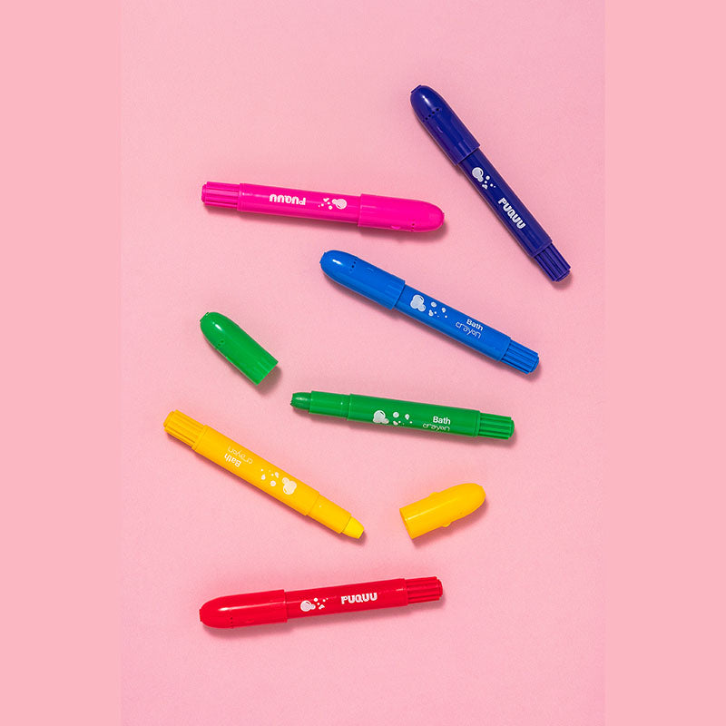 Six colorful bath crayons in pink, blue, green, yellow, and red, arranged diagonally on a solid pink background. Two green caps and two yellow caps are placed beside their respective pens.