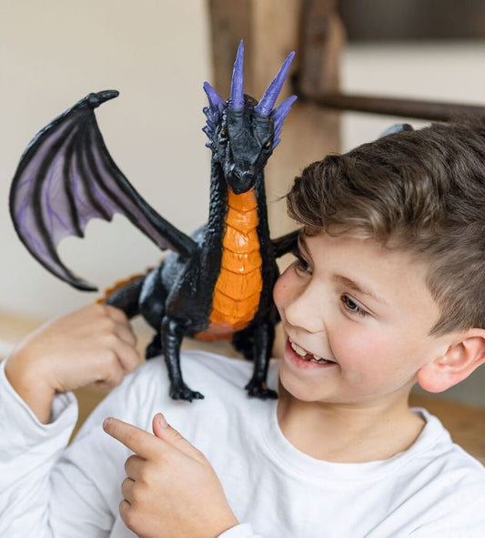 A smiling young boy in a white long-sleeve shirt holds and points to a black dragon toy with purple wings and an orange underbelly. The boy is indoors, and the dragon is positioned on his shoulder.