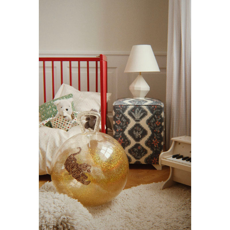 A styled children's bedroom scene with a red crib, patterned ottoman, white lamp, and a large clear glitter ball containing a tiger illustration on the floor. A plush blanket and curtains are also visible.