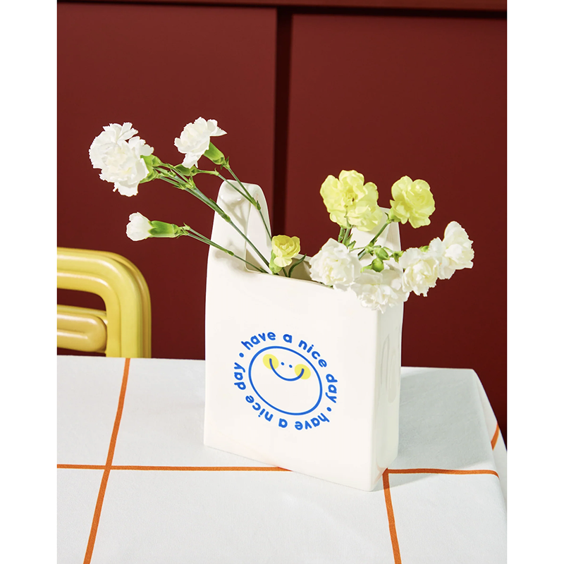 A white ceramic bag with a blue smiley face logo and the text 'have a nice day' sits on a table covered with a white and orange checkered cloth. White and pale yellow flowers are tucked into the bag, which is set against a dark red background with a yellow chair partially visible.