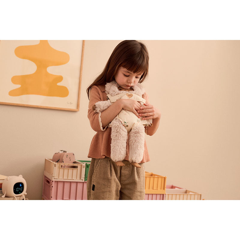 A young girl with dark hair stands in a warm-toned room holding a fluffy cream-colored plush toy. She is wearing a rust-brown long-sleeve top and brown pants.