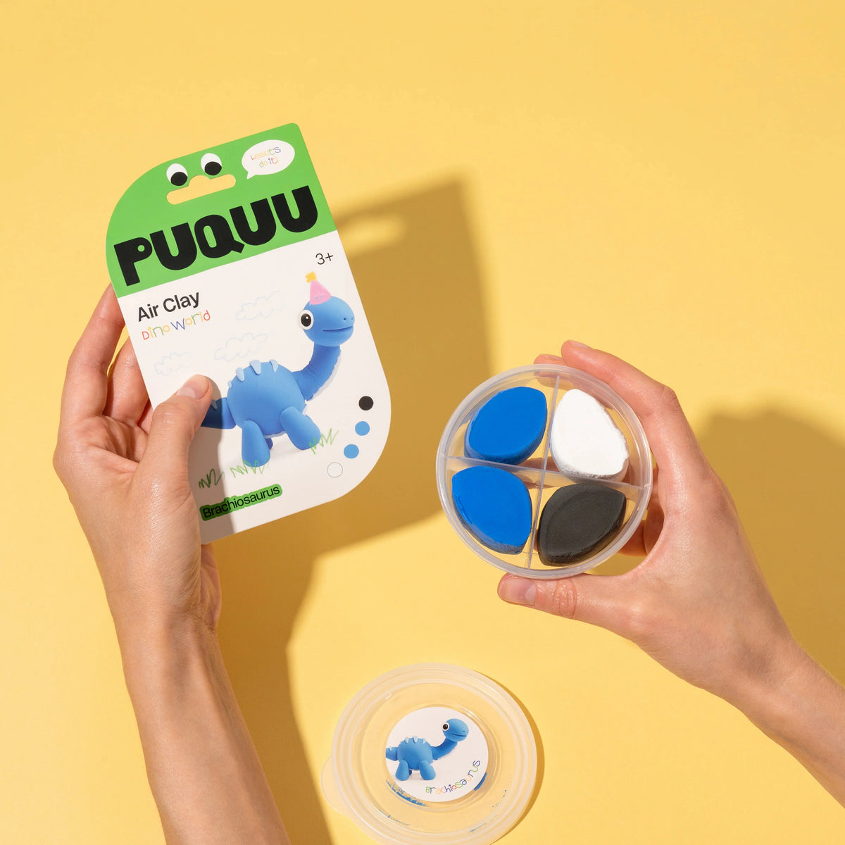 A person holds a package of PUQUUU Air Clay playdough and a round four-compartment tray showing the colored clay pieces. Another tray is shown below with a printed insert.