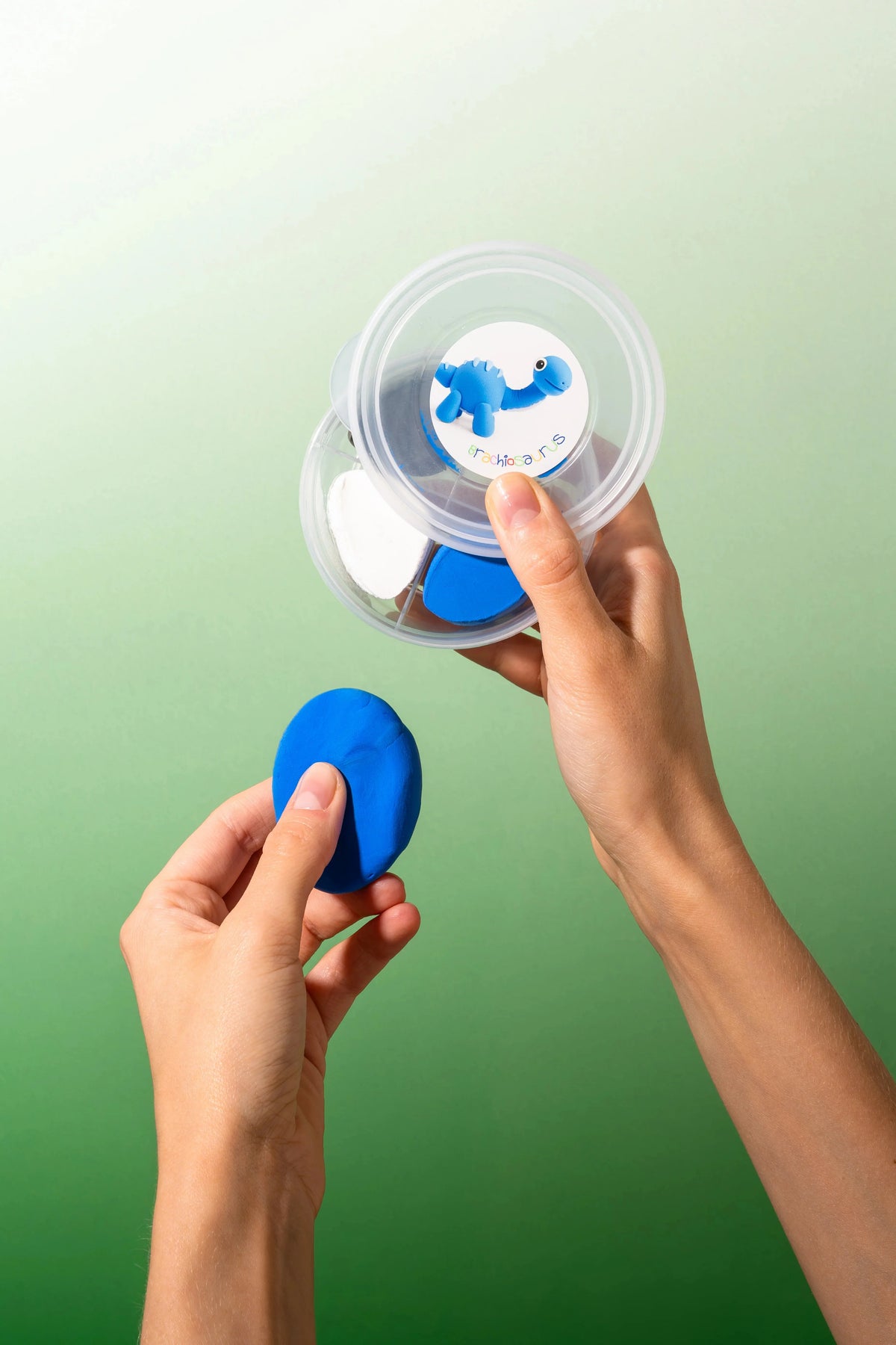 Two hands holding a small clear plastic container filled with colorful oval sponges, with one hand holding a bright blue piece of air clay.
